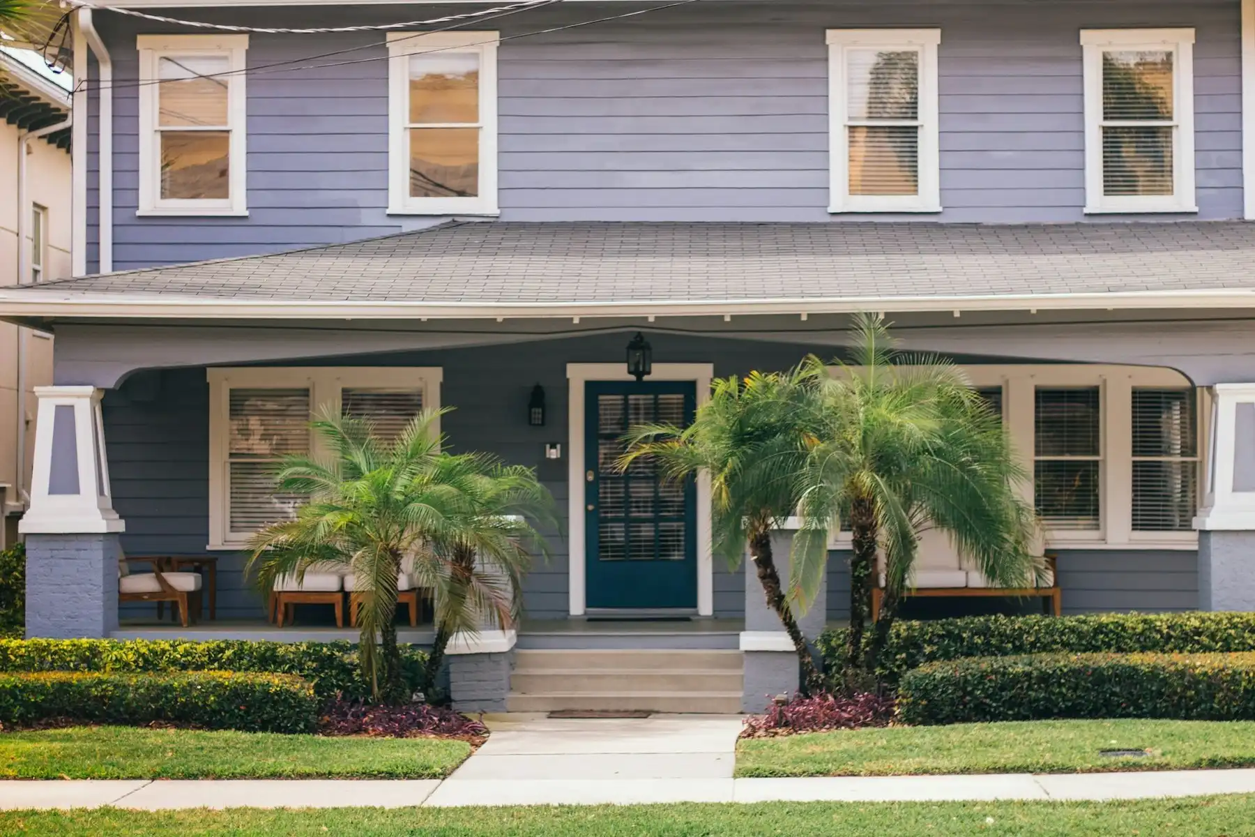 A pale blue Florida bungalow with white trim, a deep blue front door and two pygmy date palms beside the steps, on a sunny morning