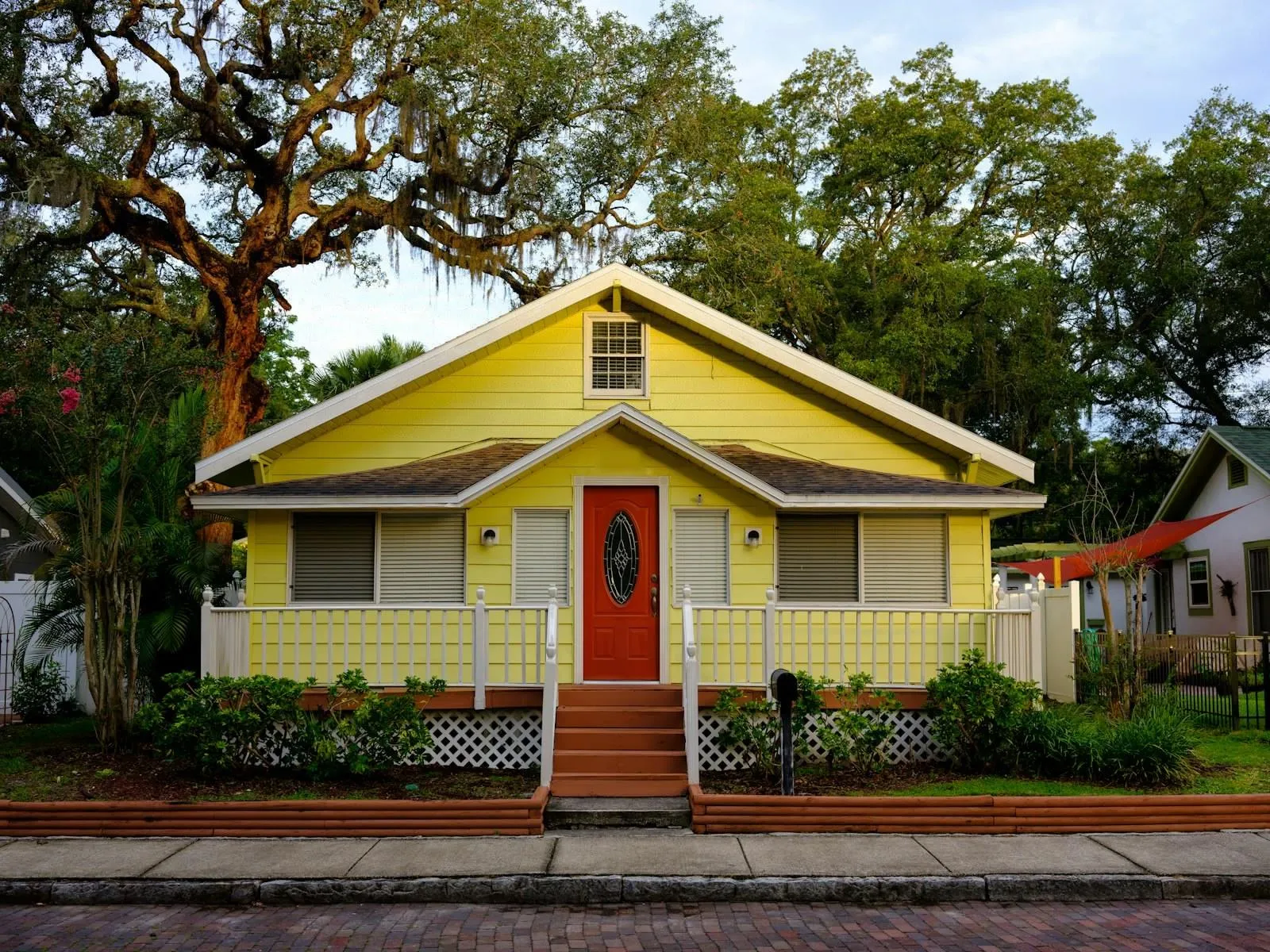 A yellow Florida bungalow with a red front door and white porch rails under large oak trees