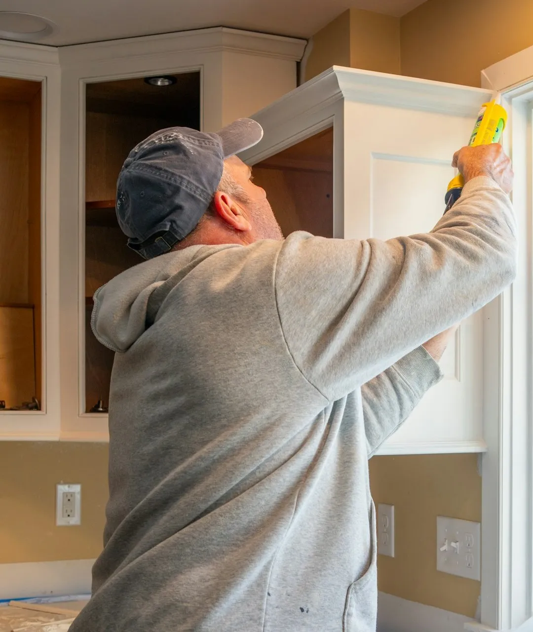 A man in a gray hoodie runs a bead of caulk along the top of a white cabinet beside a window frame