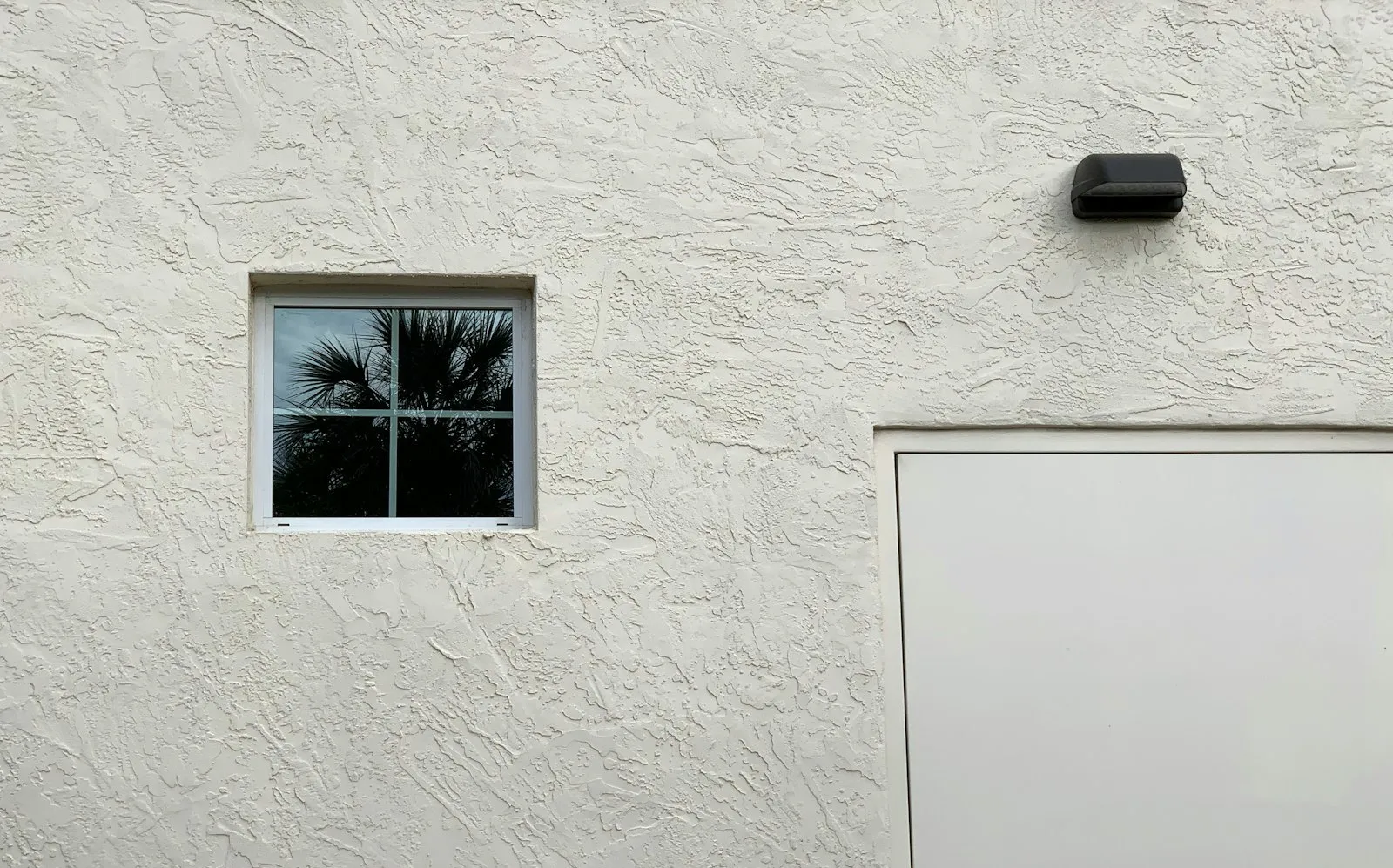 A sunny white stucco wall with a small window reflecting palm trees